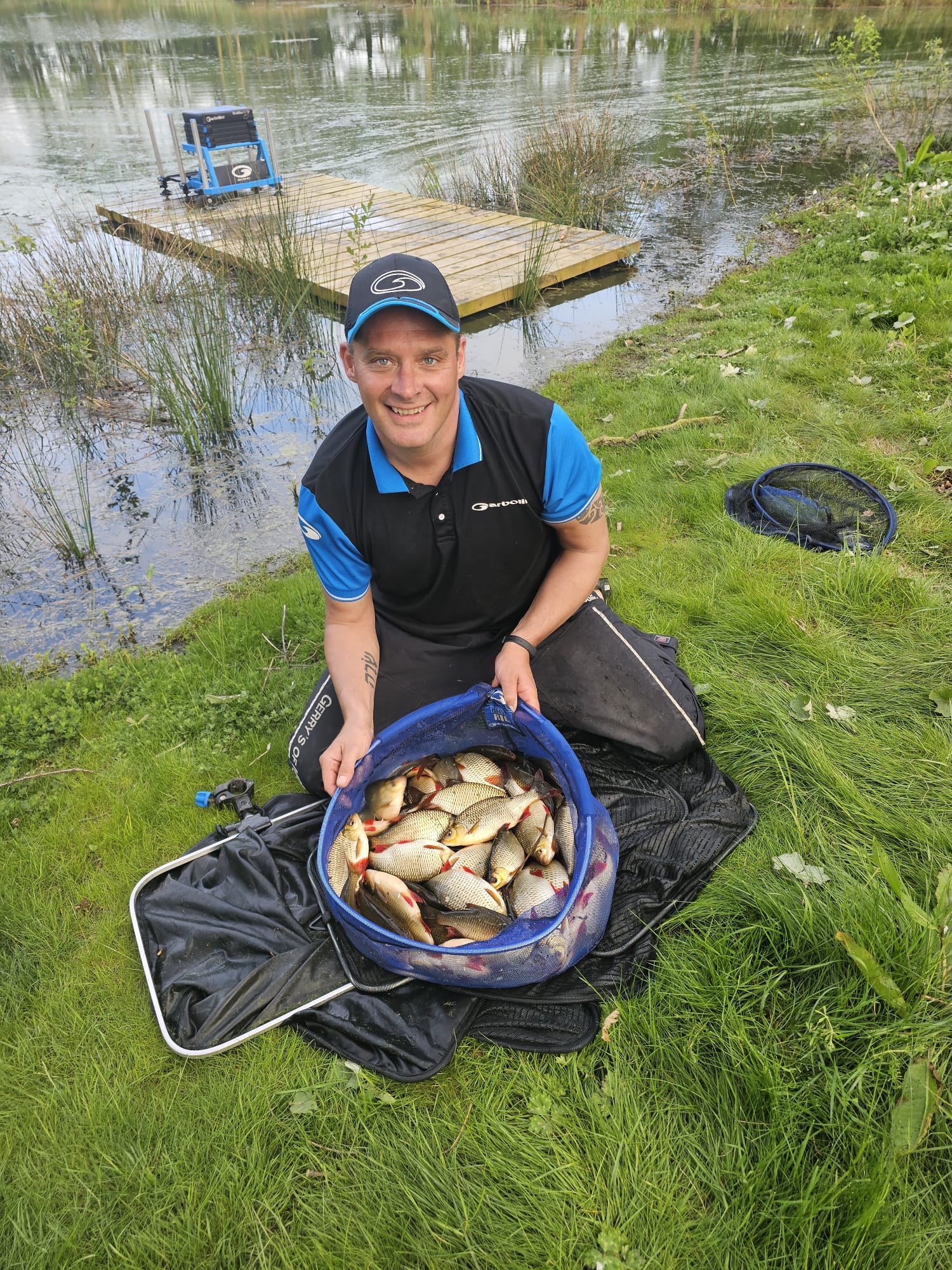 Angler smiling while holding a net filled with freshly caught fish, seated on the grass by a fishing lake with a wooden platform in the background, representing a relaxing fishing experience at Woodland Lakes Holiday Lodges.