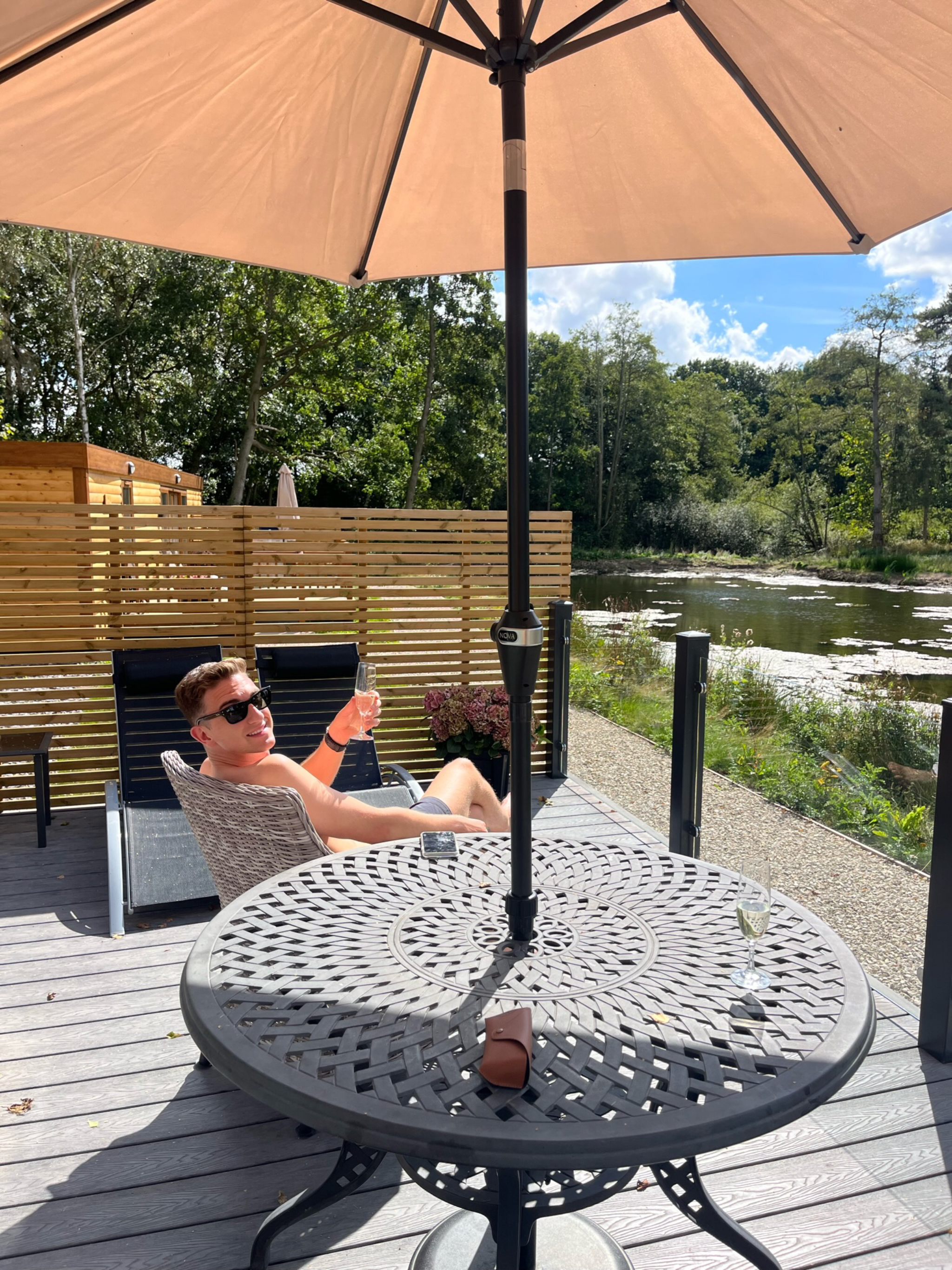 Man relaxing on a deck chair under an umbrella, enjoying a drink by a fishing lake at Woodland Lakes Holiday Lodges, surrounded by nature.
