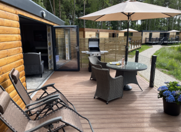 Outdoor deck area of Rowan Boutique Lodge featuring a table and chairs, barbecue grill, and sun umbrella, surrounded by lush greenery and other lodges, highlighting luxury and comfort for couples and their dogs.