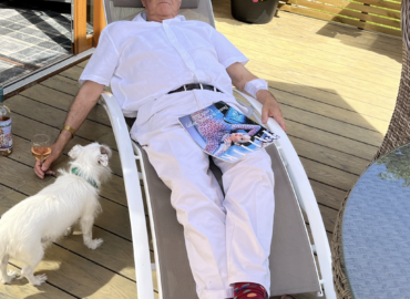 Man relaxing in white attire on a sun lounger with a dog nearby, holding a magazine and a drink, set in a peaceful outdoor lodge environment.