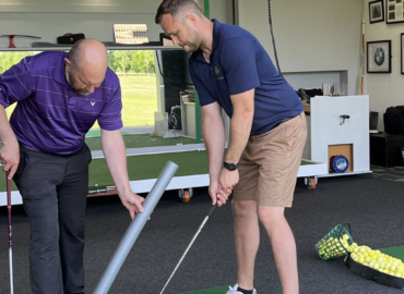 Two men engaged in a golf lesson at a driving range, one demonstrating swing technique while the other observes, with golf balls and equipment visible in the background.