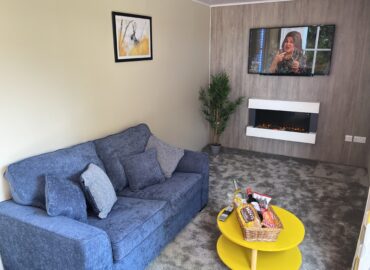 Cosy lounge area in Rowan Boutique Lodge featuring a blue sofa, yellow coffee table, wall-mounted TV, and modern electric fireplace, designed for couples seeking a romantic getaway.