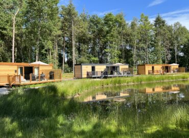 Rowan Boutique Lodges set amidst lush greenery and a tranquil pond, featuring wooden lodges with outdoor decking, ideal for couples and their dogs seeking a romantic getaway.
