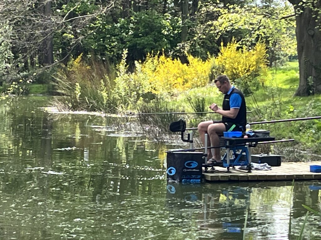 Man fishing from a wooden platform by a tranquil fishing lake, surrounded by lush greenery and reflections in the water, highlighting the serene atmosphere of Woodland Lakes holiday lodges.
