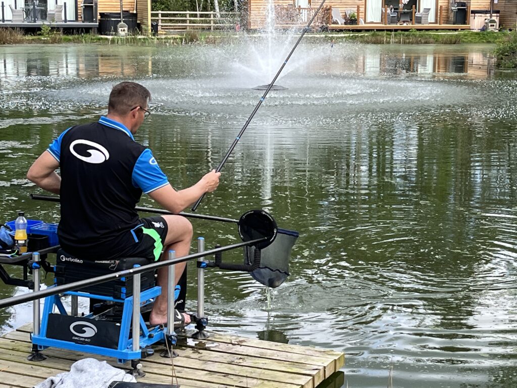 fisherman on a jetty casting out over a lake with lodges with fishing lakes in the background
