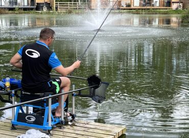 fisherman on a jetty casting out over a lake with lodges with fishing lakes in the background