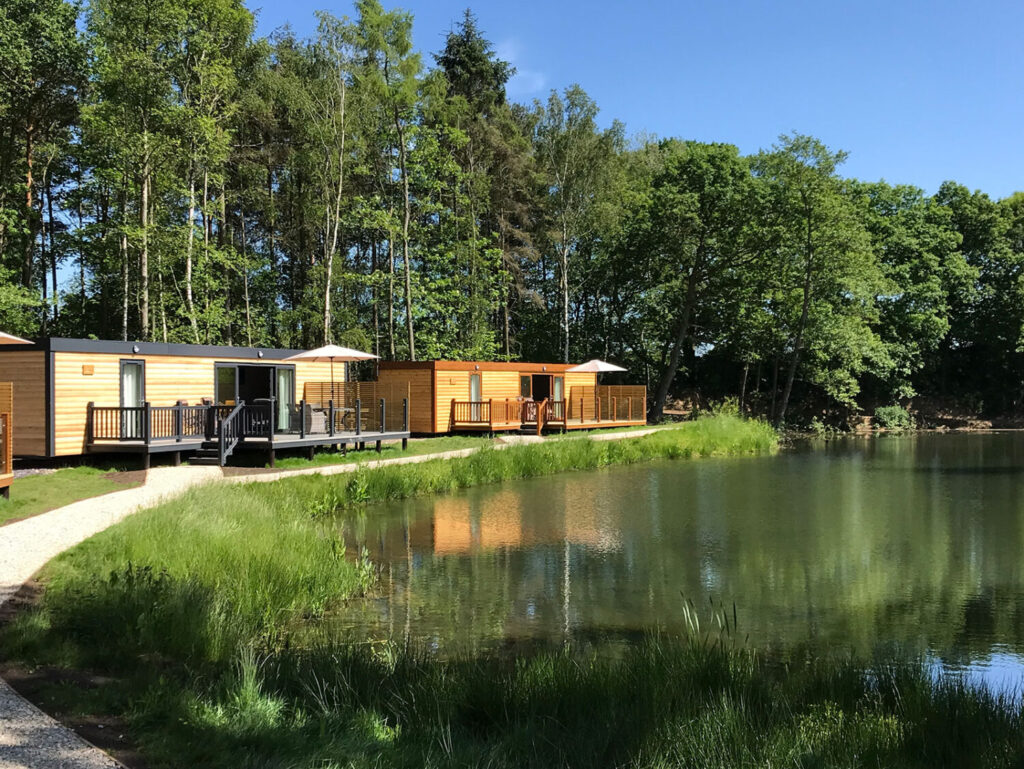 Lodges with private decks by a serene lake, surrounded by trees, showcasing luxury finishes and hot tubs for a relaxing dog-friendly getaway at Woodland Lakes Holiday Lodges.