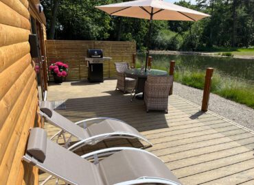 Deck area of Rowan Boutique Lodge with sun loungers, outdoor dining set, barbecue grill, and views of the lake and woodland.