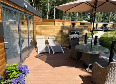 Deck area of the Rowan Boutique Lodge featuring sun loungers, a dining table, barbecue grill, and lush greenery, ideal for couples and their dogs seeking a luxury getaway.