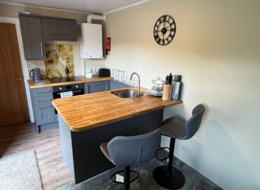 Modern kitchen area in Rowan Boutique Lodge, featuring wooden countertop, bar stools, grey cabinetry, and clock, designed for couples and dog-friendly stays.
