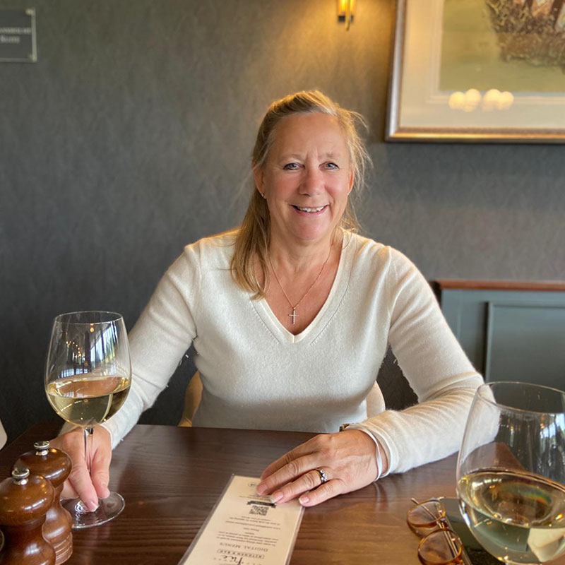 Sheila Nutt, Director of Finance at Woodland Lakes, smiling at a table with a glass of white wine and a menu, set in a relaxed dining environment.