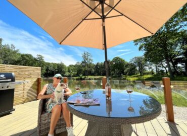 Couple enjoying drinks at a table under an umbrella, with a dog on their lap, overlooking a scenic lake and woodland at Rowan Boutique Lodge.