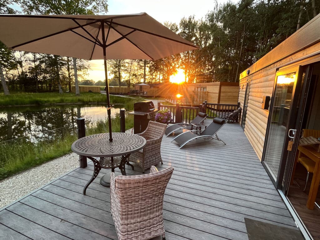 Dog-friendly lodge deck overlooking a tranquil lake at sunset, featuring outdoor seating, sun loungers, and a barbecue, ideal for couples seeking relaxation and fishing opportunities in Yorkshire.