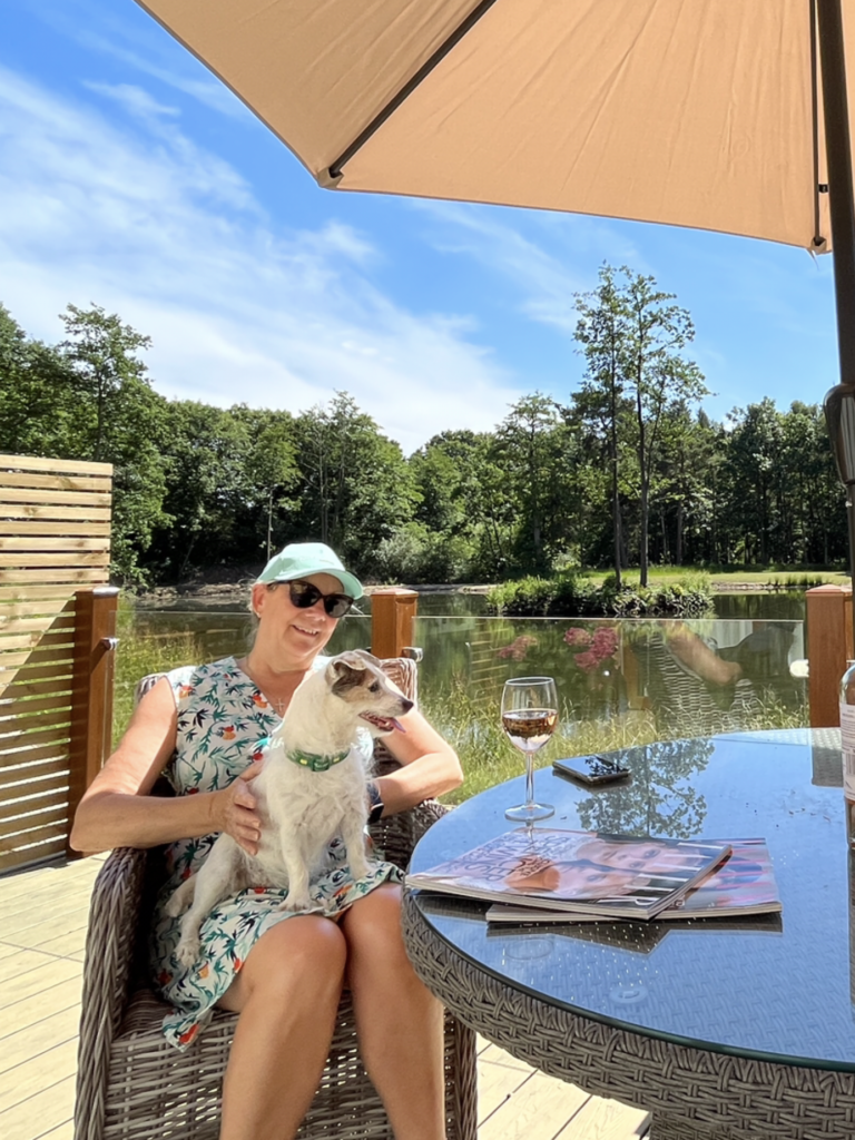 Woman relaxing in a wicker chair with a small dog on her lap, enjoying a drink at a lakeside lodge, surrounded by nature and a clear blue sky, highlighting dog-friendly accommodation at Woodland Lakes.