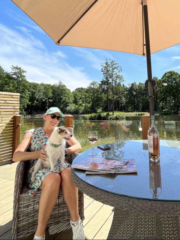 Woman sitting with a dog at a table on a sunny deck, enjoying a glass of wine, surrounded by lush greenery and a serene lake, emphasizing the dog-friendly holiday experience at Woodland Lakes.