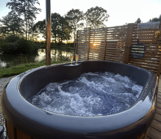 Hot tub with bubbling water and two glasses on the edge, surrounded by wooden decking and a scenic view of trees and a lake at sunset, reflecting the relaxing atmosphere of Woodland Lakes holiday lodges near York.